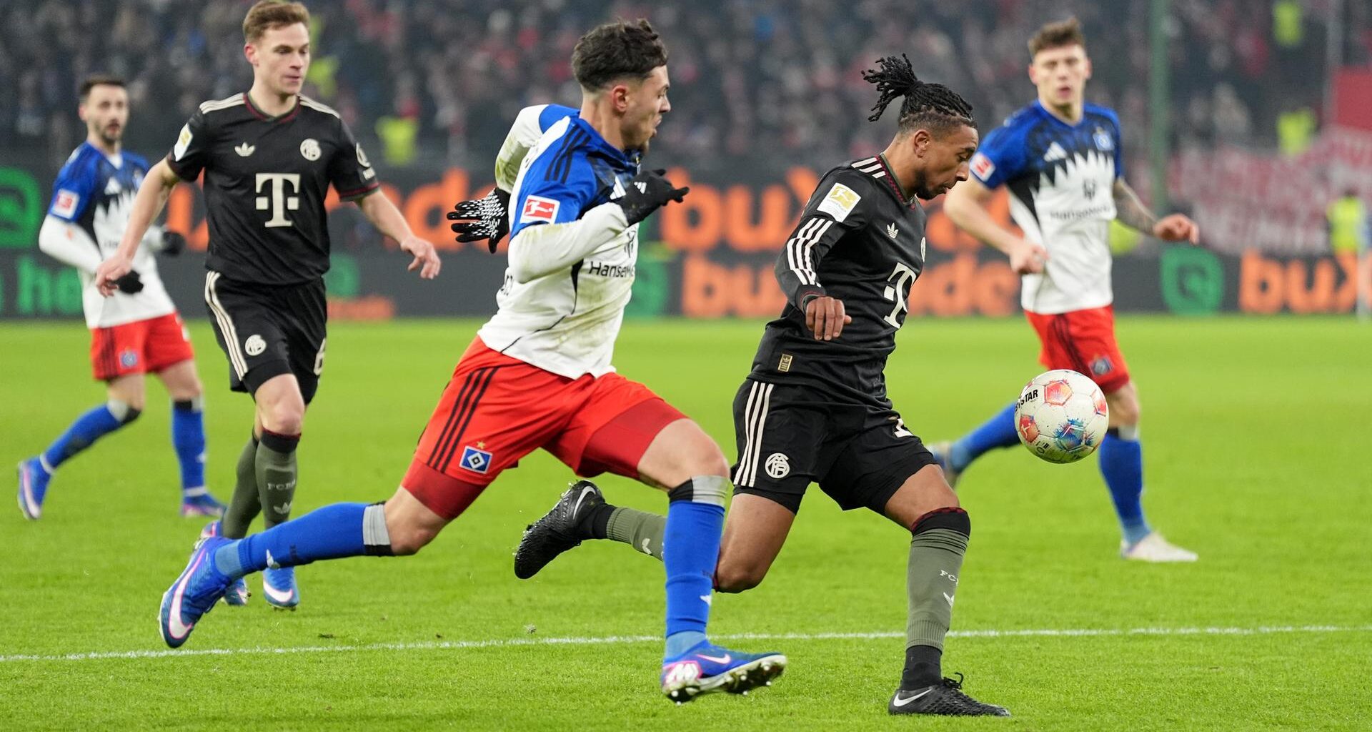 Das Bild zeigt zwei Fußball-Spieler in vollem Lauf beim Kampf um den Ball. In Hintergrund sind Mitspieler zu sehen, die das Geschehen beobachten.