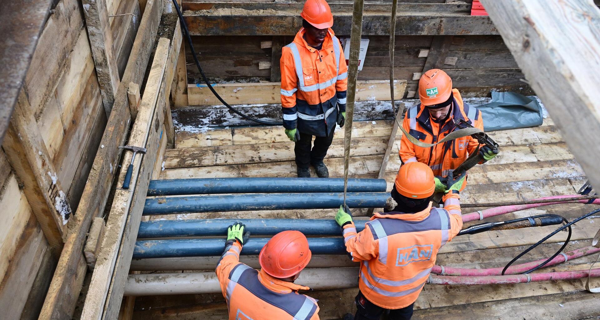 Vier Menschen mit orangefarbenen Helmen und Jacken stehen und arbeiten in einer Baugrube mit Rohren und Kabeln.