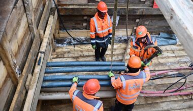 Vier Menschen mit orangefarbenen Helmen und Jacken stehen und arbeiten in einer Baugrube mit Rohren und Kabeln.