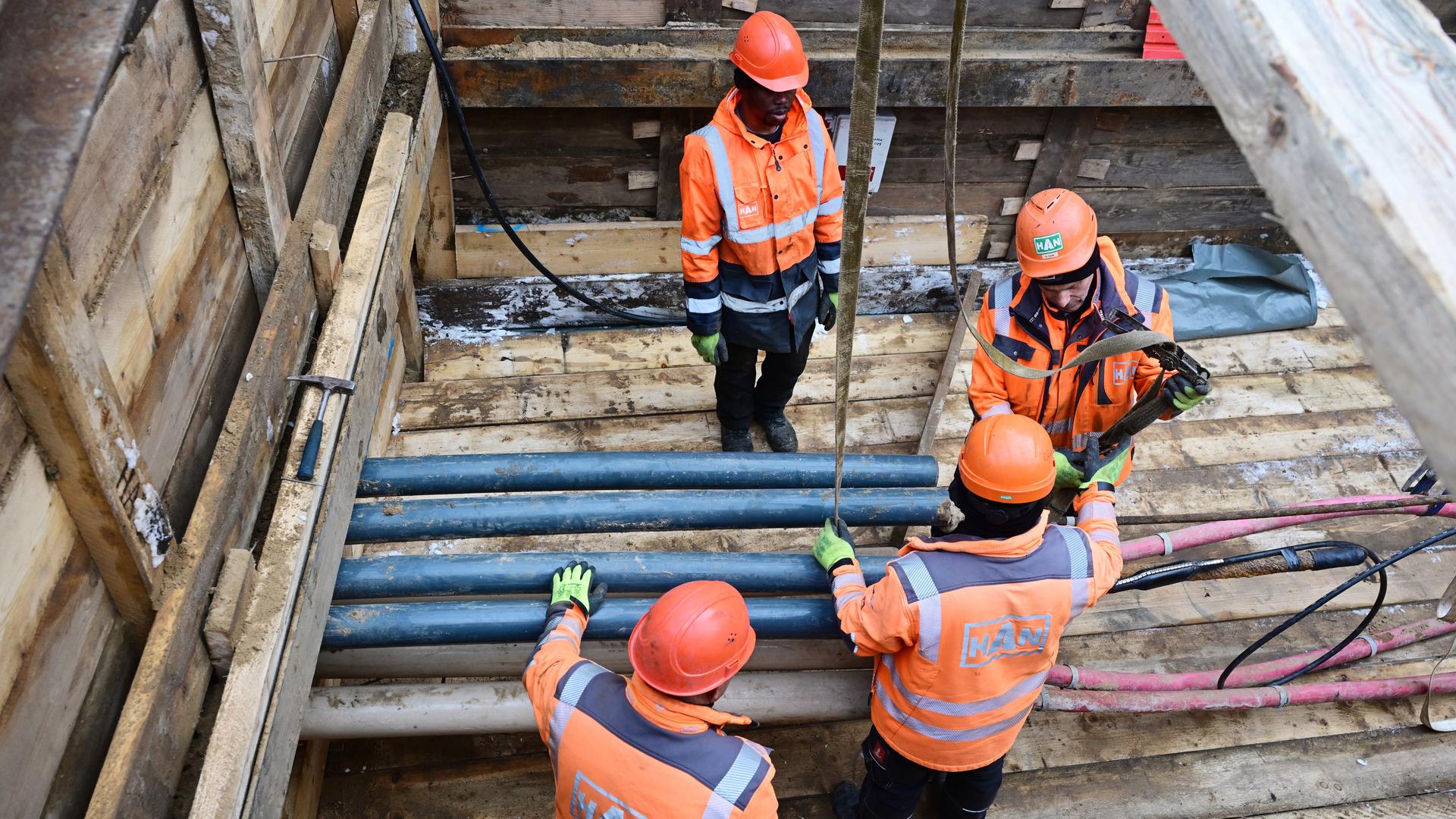Vier Menschen mit orangefarbenen Helmen und Jacken stehen und arbeiten in einer Baugrube mit Rohren und Kabeln. Vier Menschen mit orangefarbenen Helmen und Jacken stehen und arbeiten in einer Baugrube mit Rohren und Kabeln.