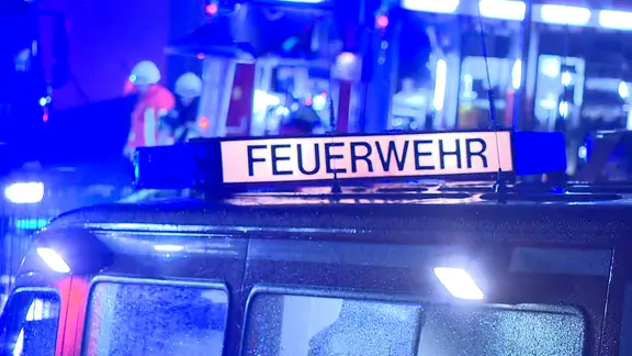 Ein Feuerwehr-Fahrzeug mit dem Schriftzug Feuerwehr steht bei Nacht vor einem Einsatzort, im Hintergrund sind Feuerwehrleute zu sehen.