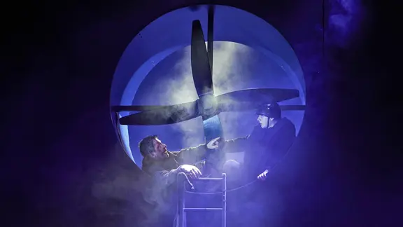 Zwei Männer sitzen in blaues Licht getaucht vor einem übergroßen Ventilator.