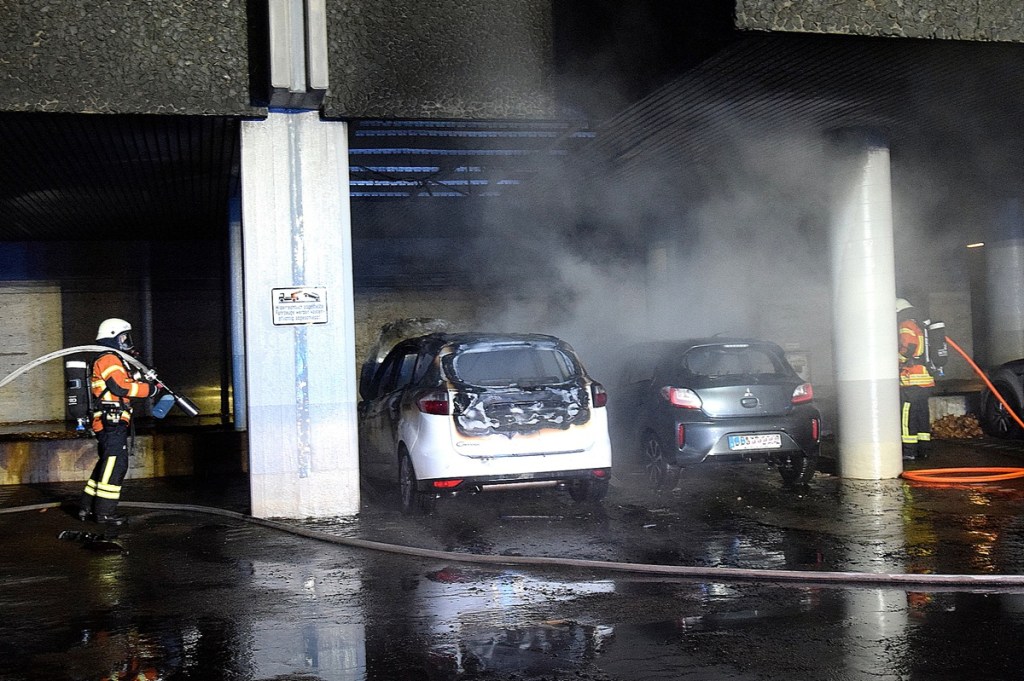 Zwei Autos brennen. Sie stehen unter dem Vordach eines Hochhauses. Einsatzkräfte der Feuerwehr löschen den Brand.
