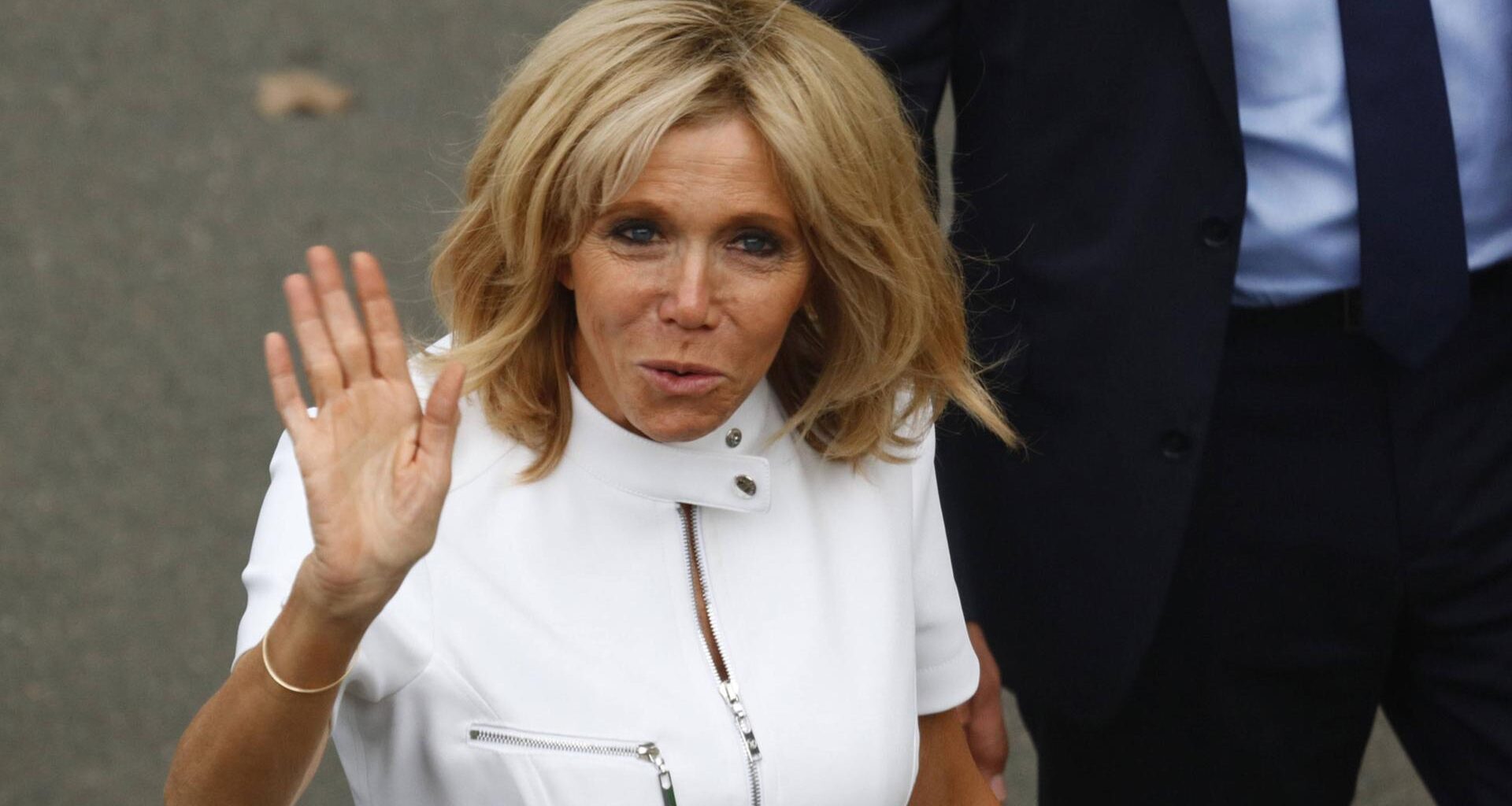Brigitte Macron im weißen Oberteil steht vor einer Wand und winkt in die Kamera.