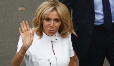 Brigitte Macron im weißen Oberteil steht vor einer Wand und winkt in die Kamera.