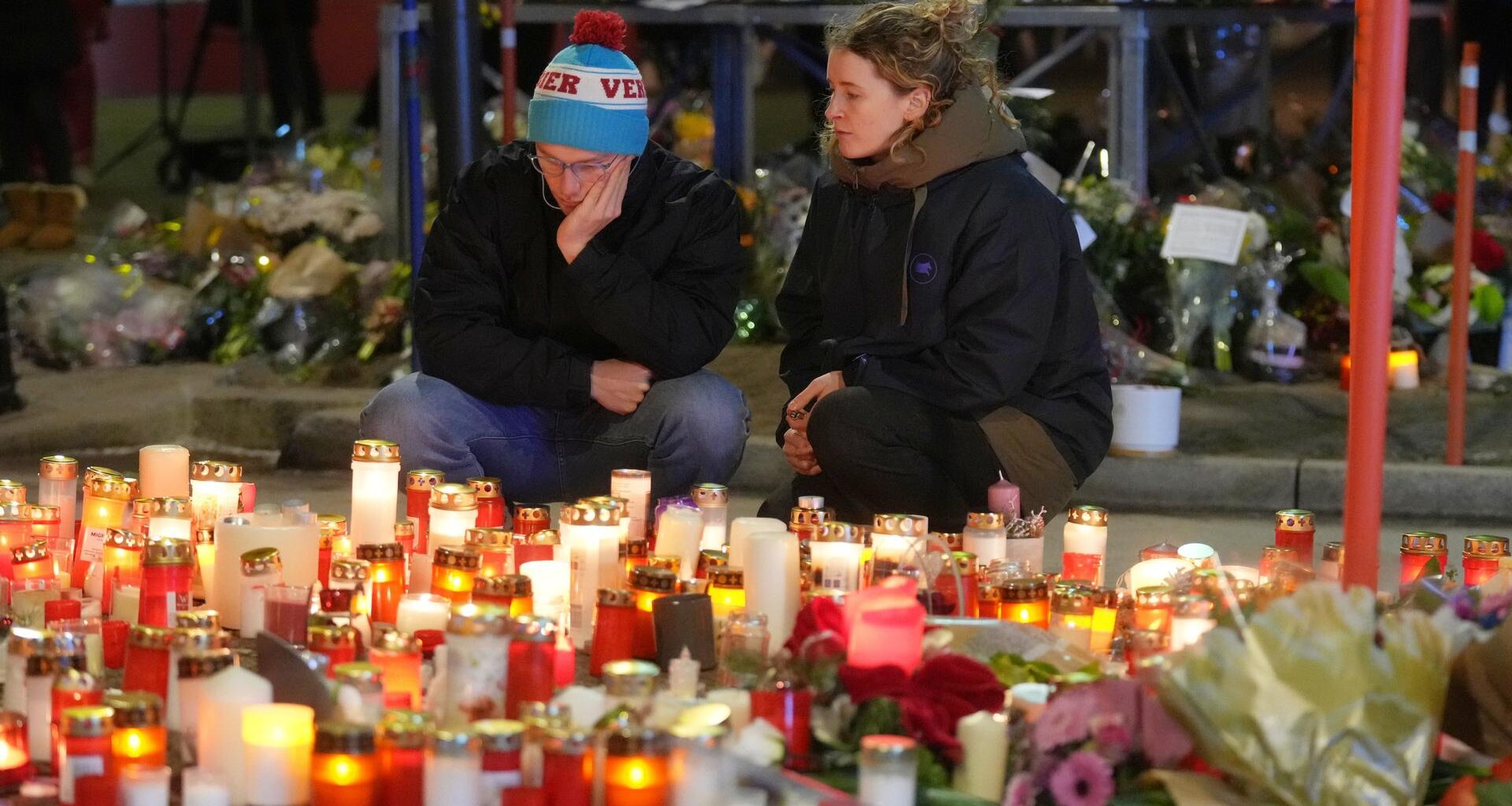 Ein Mann und eine Frau sitzen traurig vor vielen Kerzen und Blumen.