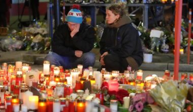 Ein Mann und eine Frau sitzen traurig vor vielen Kerzen und Blumen.