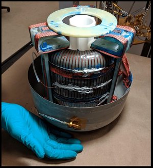 Runde Maschine, deren obere Hälfte geöffnet ist. Sie ist ungefähr so groß wie eine kleine Waschmaschine. Im Inneren befindet sich eine Mittelsäule, um die herum eine in Plastik verpackte weiße Masse enthalten ist mit kupfernen Verstebungen. Dieser Zwischenbau ist eingefasst durch mehrere äußere Begrenzungen, an deren oberem Ende längliche Platten angebracht sind.