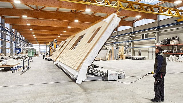 Person richtet maschinell ein vorgefertigtes Hausfassadenelement in einer Werkhalle auf.