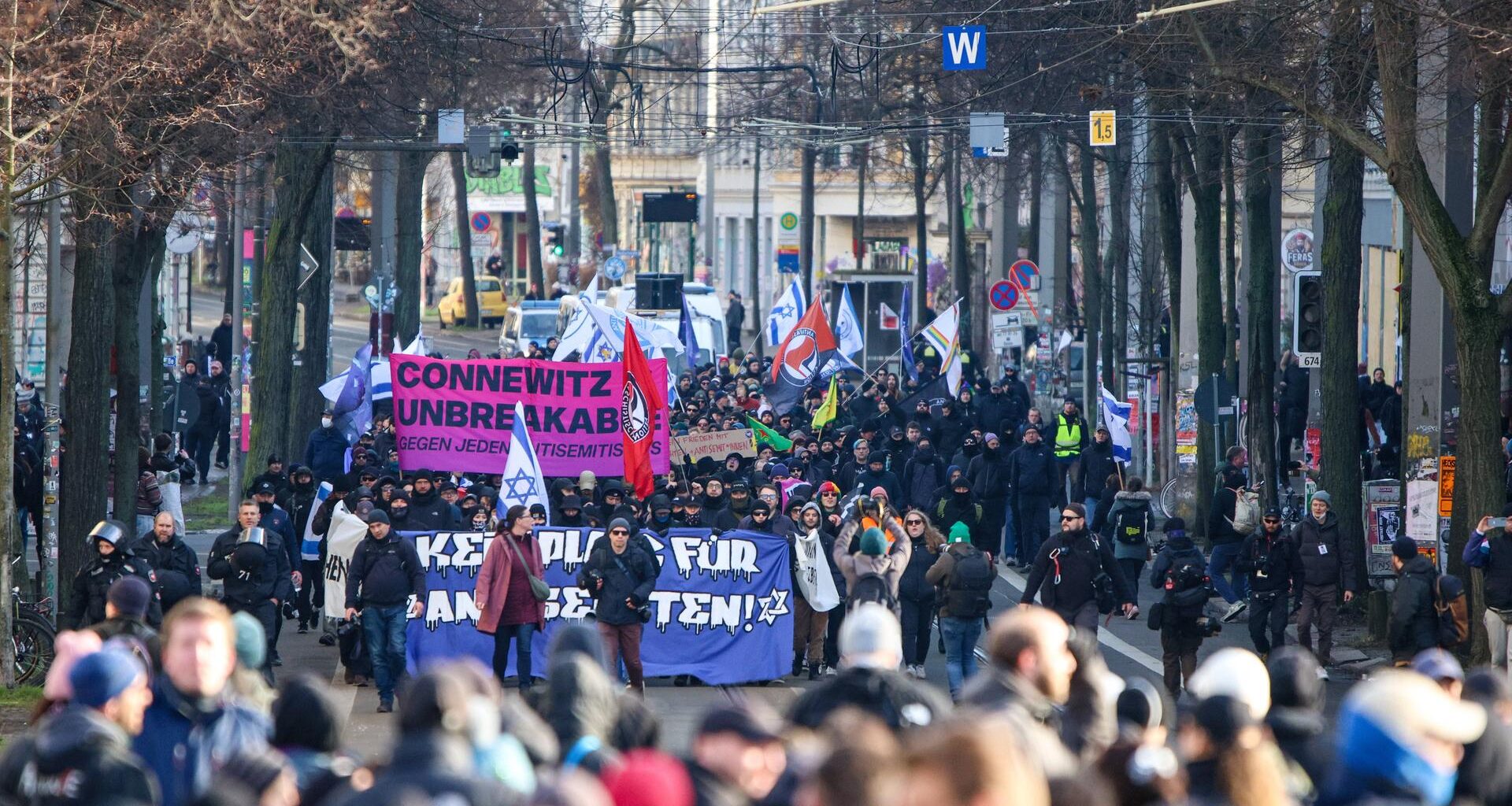 Auf einer Straße stehen sehr viele Menschen, einige tragen israelische Flaggen und Banner. Am Rand stehen viele Polizisten.