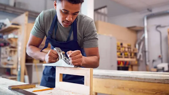 Ein Lehrling hobelt an einem Holzwerkstück.