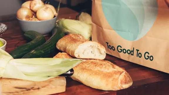 Gemüse und Baguette liegen in einer Küche, daneben eine Papiertüte mit dem Logo der Organisation "Too Good To Go". | Too Good To Go Gemüse und Baguette liegen in einer Küche, daneben eine Papiertüte mit dem Logo der Organisation "Too Good To Go".