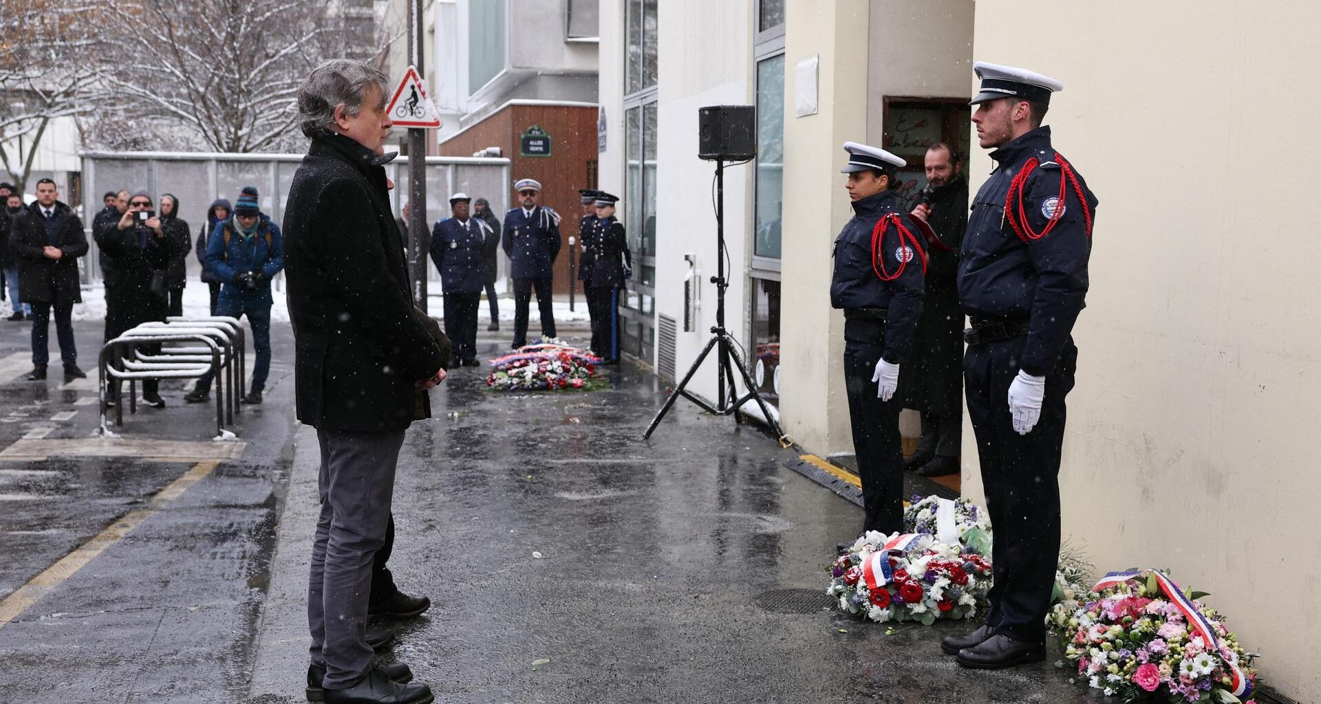 Der französische Karikaturist gedenkt der Opfer des Terroranschlags auf Charlie Hebdo. Vor ihm stehen zwei Polizisten. Auf dem Boden liegen Kränze.