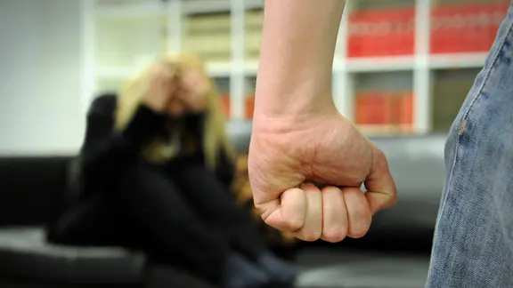 Ein Mann bedroht eine auf dem Sofa zusammengekauert sitzende Frau mit seiner Faust. (Themenbild)
