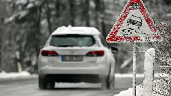 Ein Auto fährt über eine vereiste Straße, am Straßenrand warnt ein Verkehrsschild vor Glätte. (Themenbild) | Federico Gambarini/dpa Ein Auto fährt über eine vereiste Straße, am Straßenrand warnt ein Verkehrsschild vor Glätte. (Themenbild)