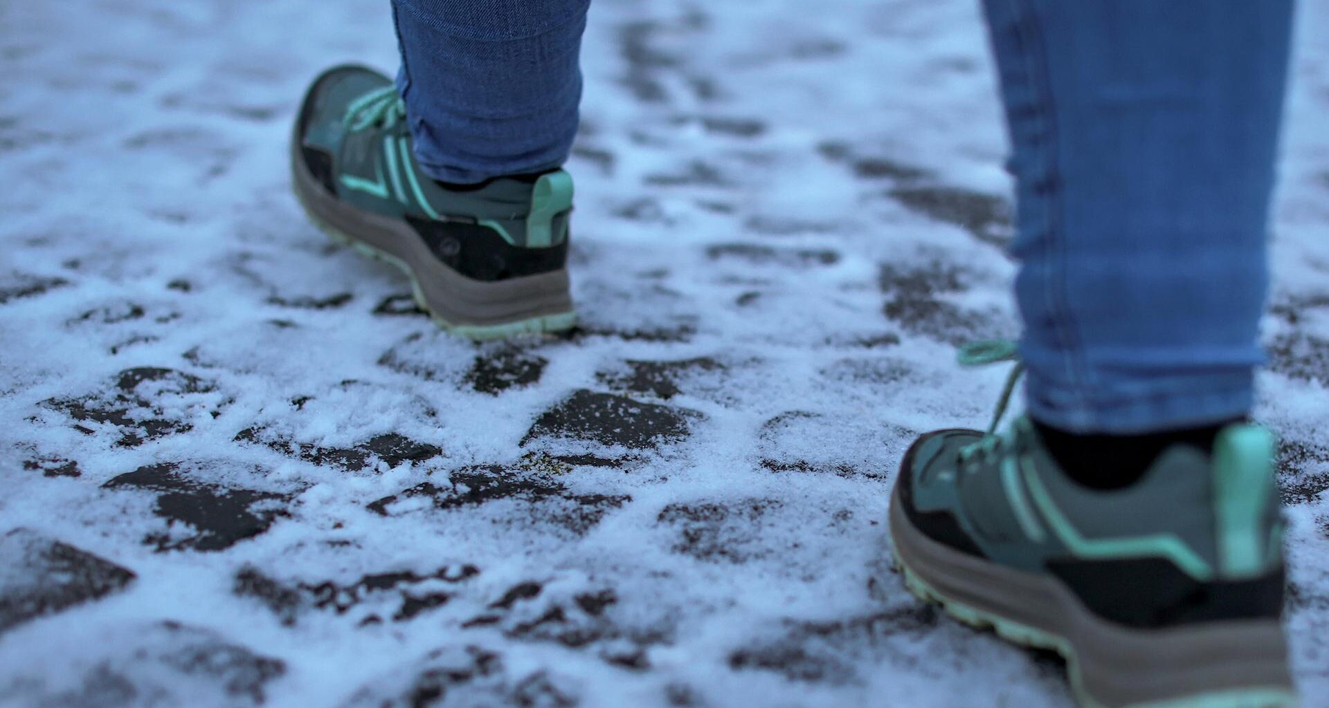 Das Bild zeigt die Nahaufnahme eines frostigen, gepflasterten Bodens und zwei Füße in Turnschuhen, die sich vorsichtig darüber bewegen.