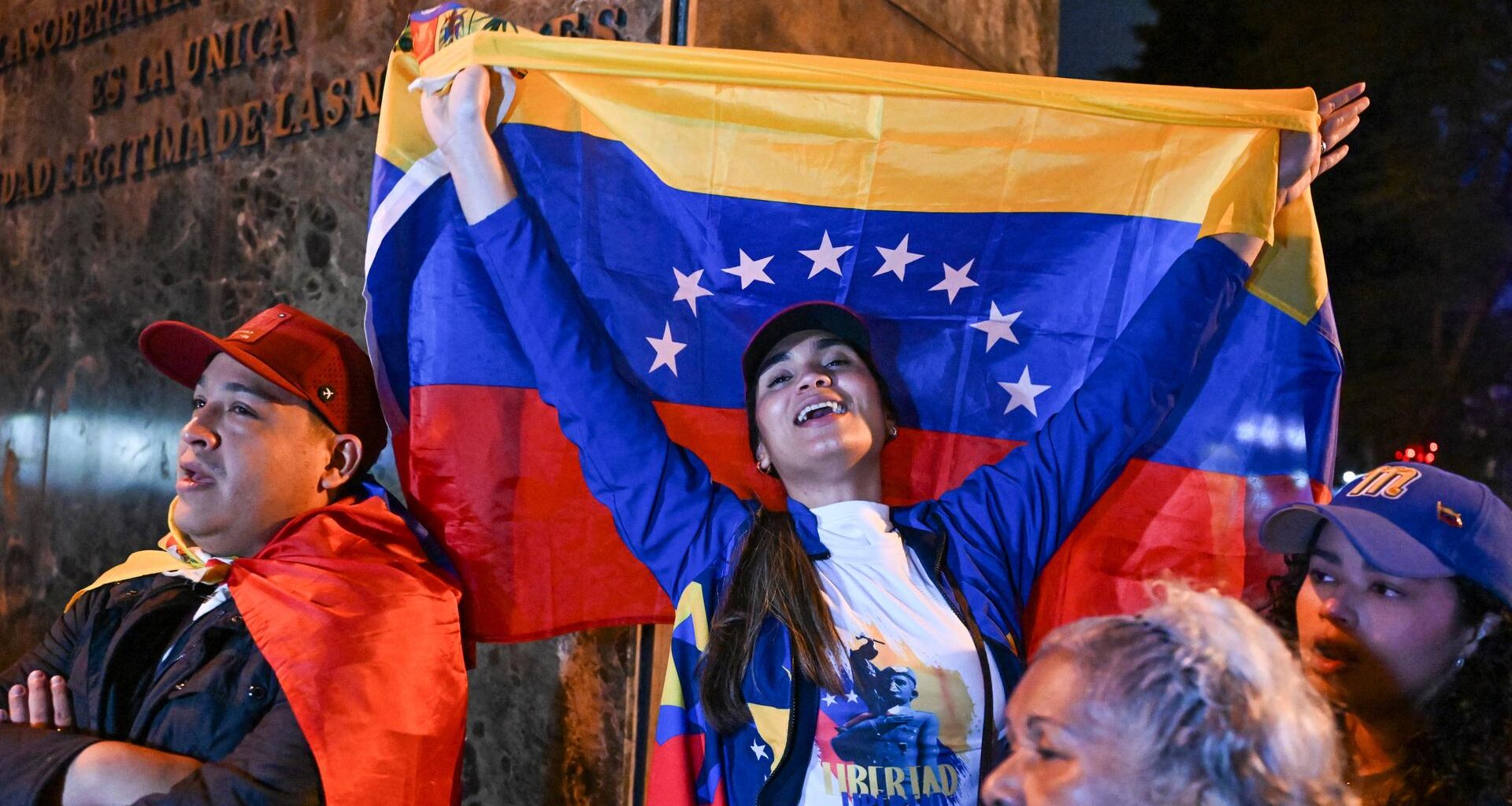 Eine Frau reißt eine venezolanische Nationalflagge in die Höhe. Sie steht zwischen weiteren Teilnehmern einer spontanen Versammlung von Exil-Venezolanern.