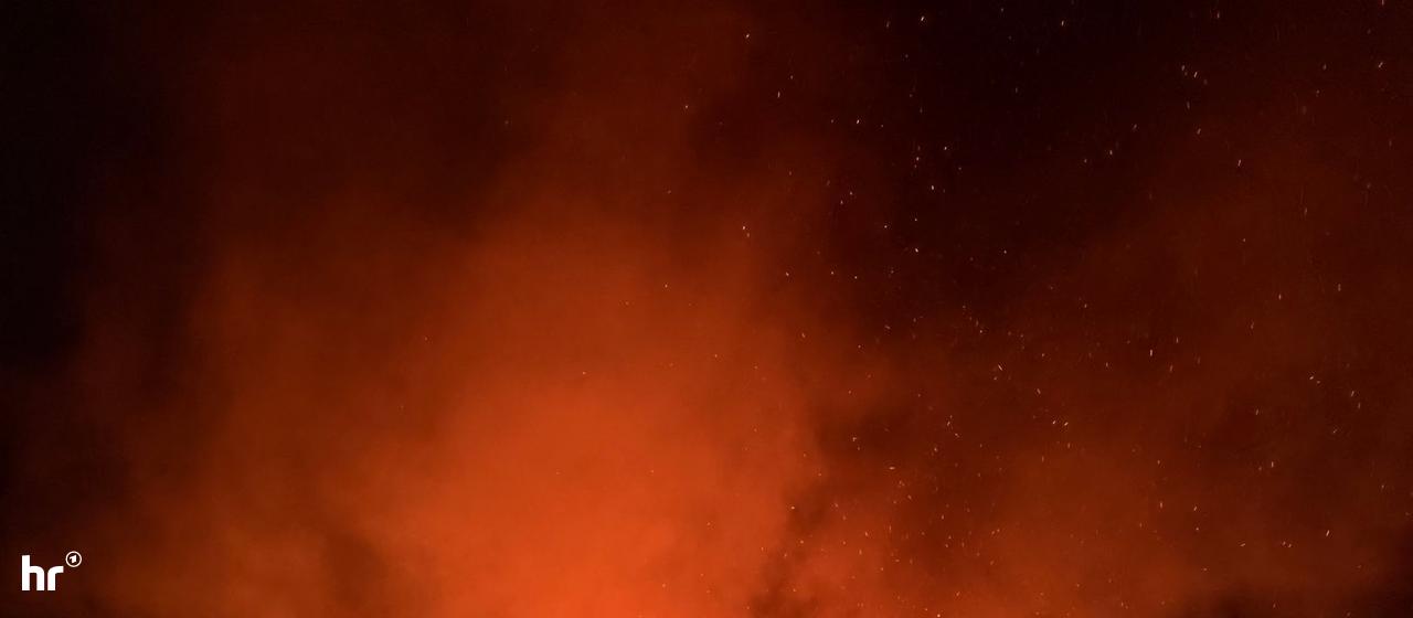 Vor einem nächtlichen Himmel ist ein lichterloh brennendes Haus auf einem Hügel in einem kleinen Ort zu sehen.