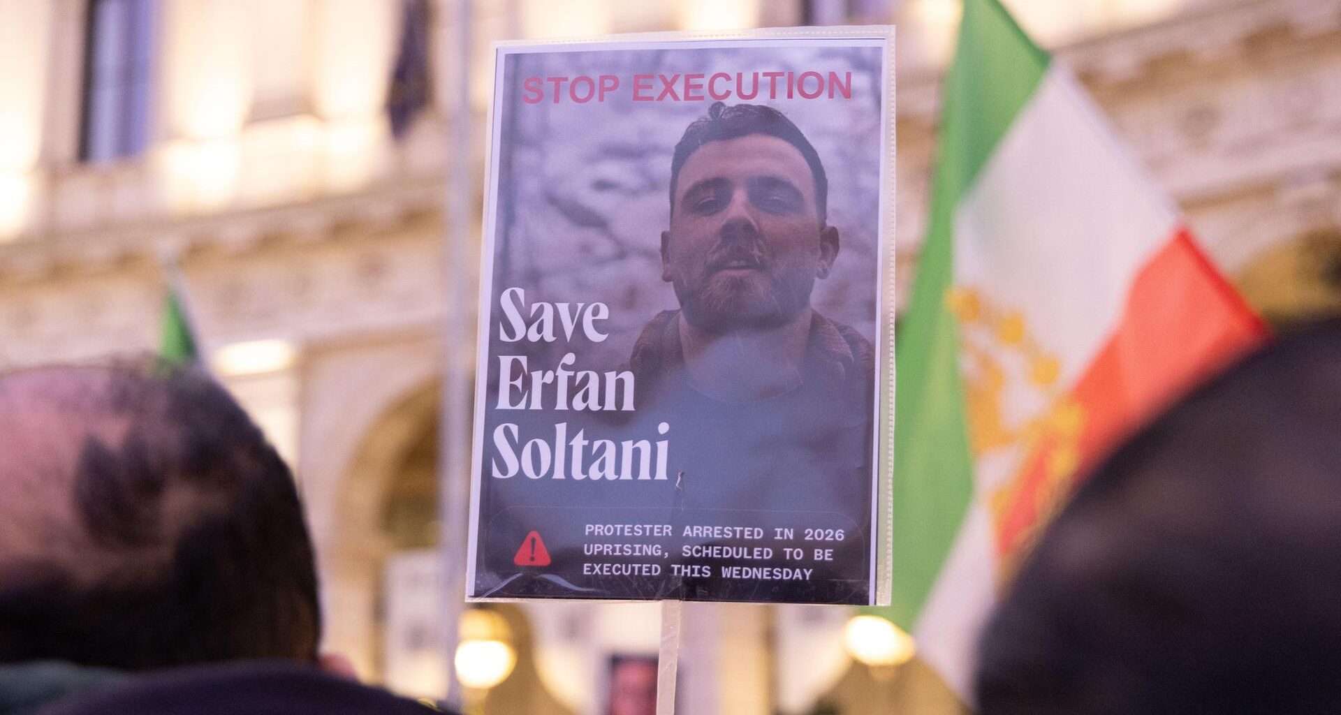 Das Bild wird mit einem Stock hochgehalten, auf dem der Oberkörper Soltanis zu sehen ist. Dahinter unscharf eine iranische Fahne und eine Gebäudefront. Im Vordergrund ebenfalls unscharf die Hinterköpfe von zwei Demonstranten.