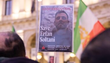 Das Bild wird mit einem Stock hochgehalten, auf dem der Oberkörper Soltanis zu sehen ist. Dahinter unscharf eine iranische Fahne und eine Gebäudefront. Im Vordergrund ebenfalls unscharf die Hinterköpfe von zwei Demonstranten.