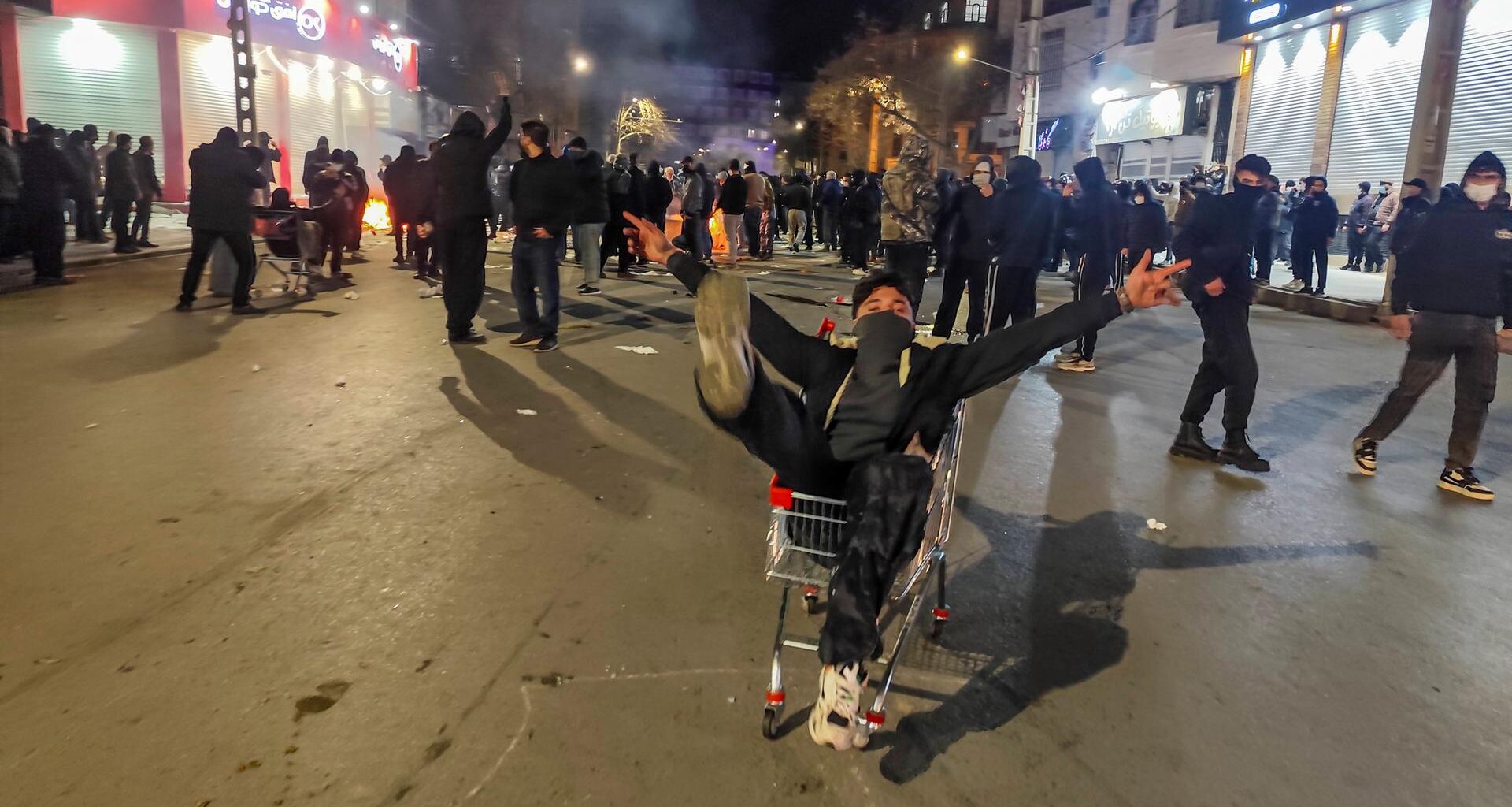 Das Bild zeigt viele Demonstranten im Dunkeln in einer Fußgängerzone. Im Vordergrund sitzt ein maskierter junger Mann in einem Einkaufswagen und steckt alle Arme und Beine von sich.