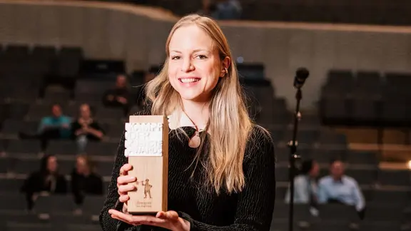 Julia Engelmann erhielt den Ehrenpreis beim Deutschen Poetry Slam Award 2026. | Michelle Jekel Julia Engelmann präsentiert lächelnd eine hölzerne Trophäe in die Kamera.