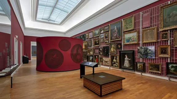 Ein Ausstellungsraum mit roten Wänden und Bildern, die an einem weißen Gitternetz hängen