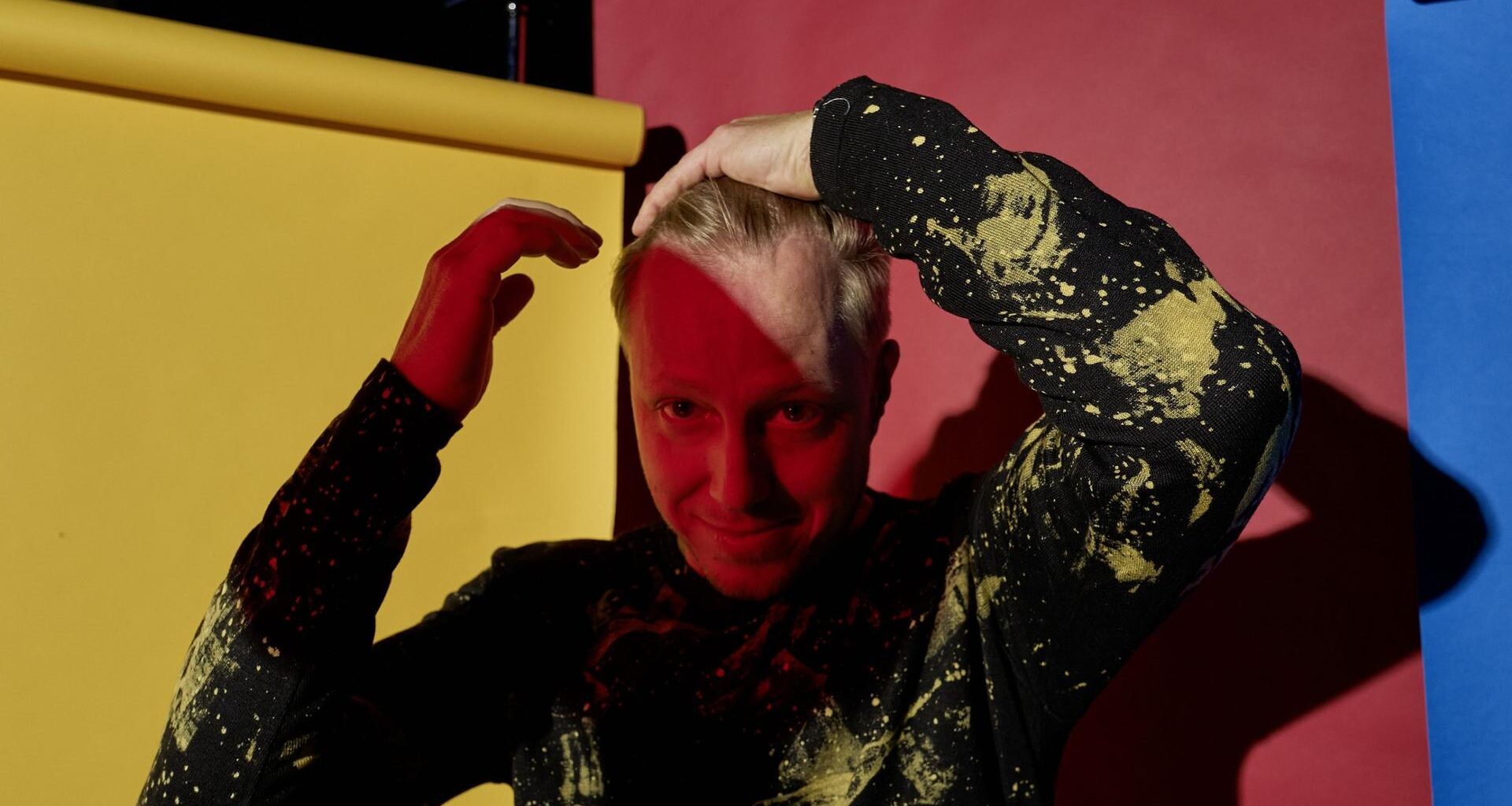 Ein buntes Portrait vor Papierbahnen in rot, gelb und blau. Der Musiker Martin Tingvall streicht sich die blonden Haare zurück, er trägt einen schwarzen Pullover mit wildem, gelbem Muster. Er ist halb im Schatten.