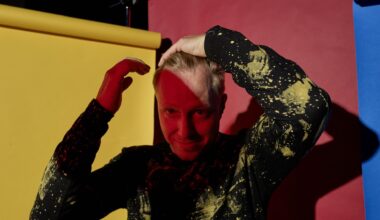 Ein buntes Portrait vor Papierbahnen in rot, gelb und blau. Der Musiker Martin Tingvall streicht sich die blonden Haare zurück, er trägt einen schwarzen Pullover mit wildem, gelbem Muster. Er ist halb im Schatten.