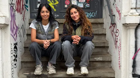 Muschda Sherzada und Gülay Ulaş sitzen auf den Stufen einer Treppe im Hauseingang