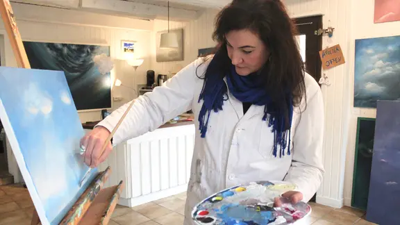 Eine Frau mit langen dunklen Haaren und weißem Kittel bemalt mit einem Pinsel eine Leinwand mit einer Wolkenlandschaft | Elvira Vogeler Eine Frau mit langen dunklen Haaren und weißem Kittel bemalt mit einem Pinsel eine Leinwand mit einer Wolkenlandschaft