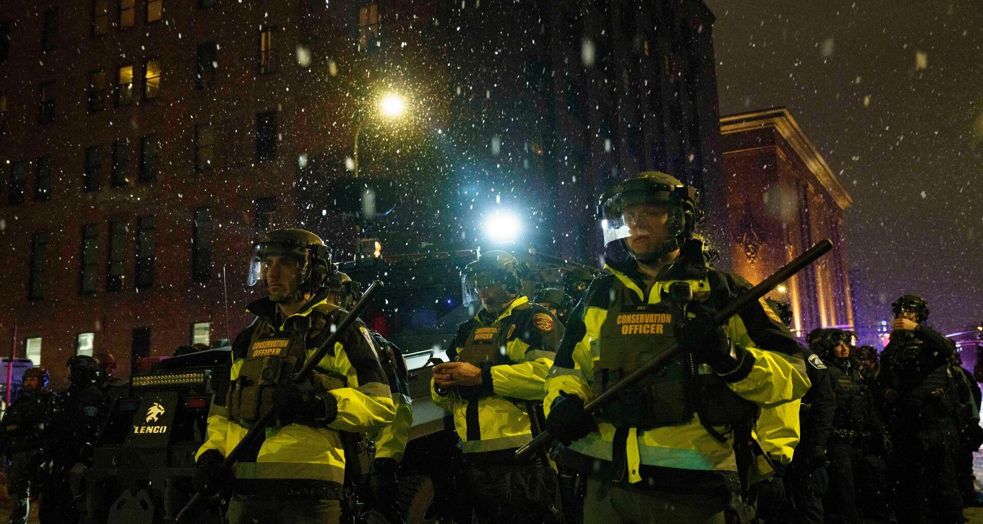 Polizeibeamte stehen auf einer Staße in Minneapolis. Sie tragen Schlagstöcke. Es ist dunkel und schneit.