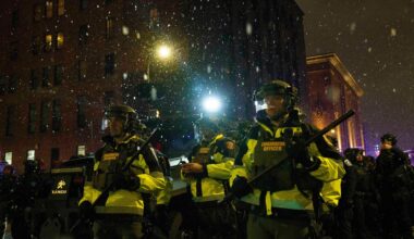 Polizeibeamte stehen auf einer Staße in Minneapolis. Sie tragen Schlagstöcke. Es ist dunkel und schneit.