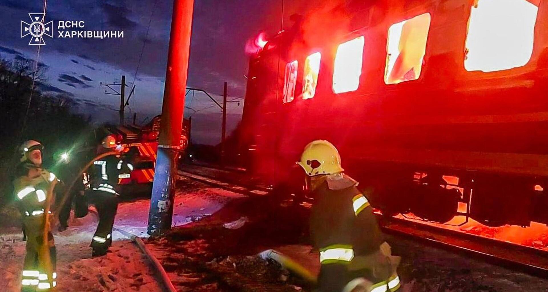 An einer Gleisstrecke legen mehrere Feuerwehrleute einen Schlauch. Im Hintergrund schlagen Flammen aus einem Zugwaggon.