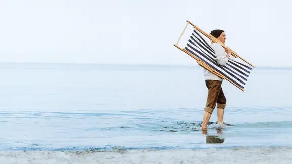 Ein Mann läuft mit einer zusammengeklappten Sonnenliege über der Schulter durch das Wasser.