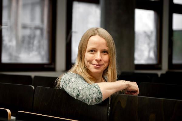 Professorin Annabelle Bohrdt sitzt lächelnd zwischen Reihen dunkler Hörsaalstühle, natürliches Licht fällt durch große Fenster im Hintergrund.