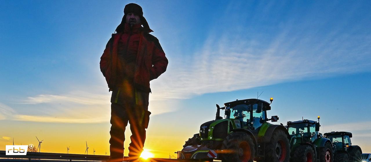 08.01.2024, Brandenburg, Jacobsdorf: Landwirte blockieren am frühen Morgen mit ihren Traktoren die Auffahrt auf die Autobahn A12 in Richtung Berlin, ein Mann steht dabei im Gegenlicht der aufgehenden Sonne.(Quelle:dpa/P.Pleul)
