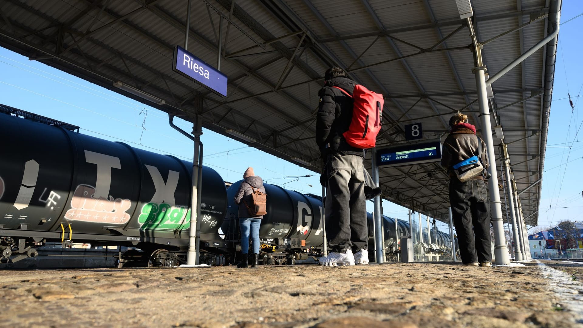 Reisende stehen auf dem Bahnsteig am Bahnhof vor einem Güterzug (Archivbild): Auf der Strecke zwischen Leipzig und Dresden kommt es am Dienstagmorgen zu erheblichen Beeinträchtigungen.