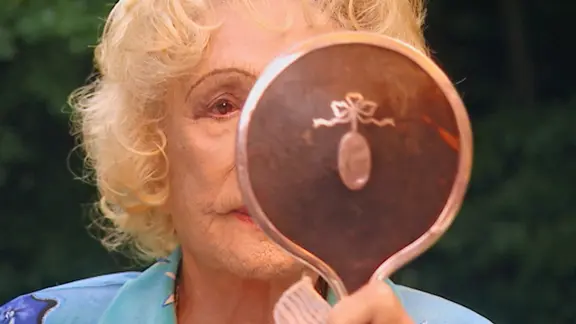 Leni Riefenstahl kontrolliert ihr Aussehen für die Aufzeichnung zur dreiteiligen Dokumentation "Speer und er" von Regisseur Heinrich Breloer (1999) | Bavaria Media Eine blonde ältere Frau ist halb verdeckt von einem Handspiegel und betrachtet ihr Spiegelbild - Lenie Riefenstahl in einer