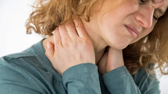 Eine Frau mit schmerzverzerrtem Gesicht hält sich mit den Händen den Nacken.