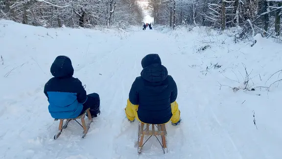 Zwei Kinder sitzen auf Schlitten auf einem verschneiten Waldweg.