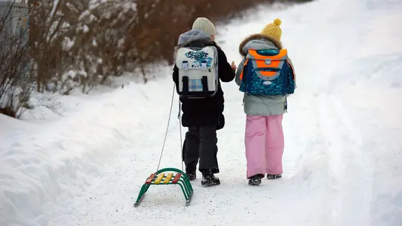 Zwei Kinder mit einem Schlitten laufen über einen mit Schnee bedeckten Gehweg.