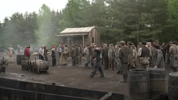 Komparsen stehen bei den Dreharbeiten zur Serie "Schwarzes Gold" auf einem Platz zwischen Ölfässern und einem Holzschuppen. | NDR Komparsen stehen bei den Dreharbeiten zur Serie "Schwarzes Gold" auf einem Platz zwischen Ölfässern und einem Holzschuppen.