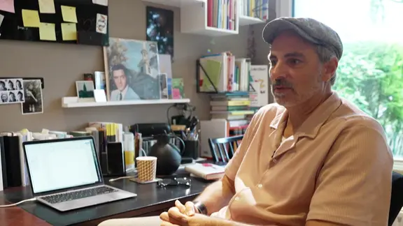 Ein Mann mit Schiebermütze sitzt am Schreibtisch in einem Arbeitszimmer. | Screenshot Ein Mann mit Schiebermütze sitzt am Schreibtisch in einem Arbeitszimmer.