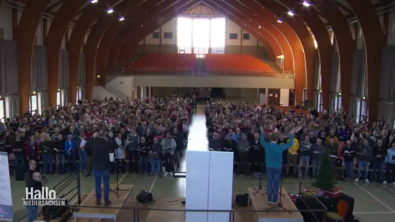 Ein Chor mit 1000 Sänger:innen bei den Proben in einer großen Halle.