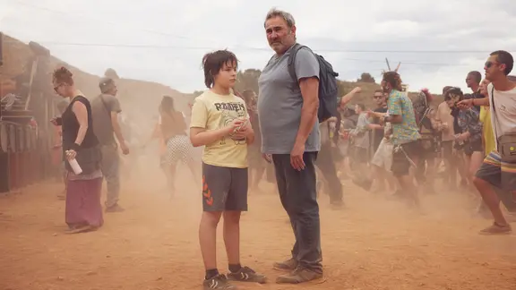 Szene aus dem Film "Sirāt": Ein Mann mit Rucksack und ein kleiner Junge stehen inmitten von tanzenden Menschen | Pandora Film / QuimVives Szene aus dem Film "Sirāt": Ein Mann mit Rucksack und ein kleiner Junge stehen inmitten von tanzenden Menschen