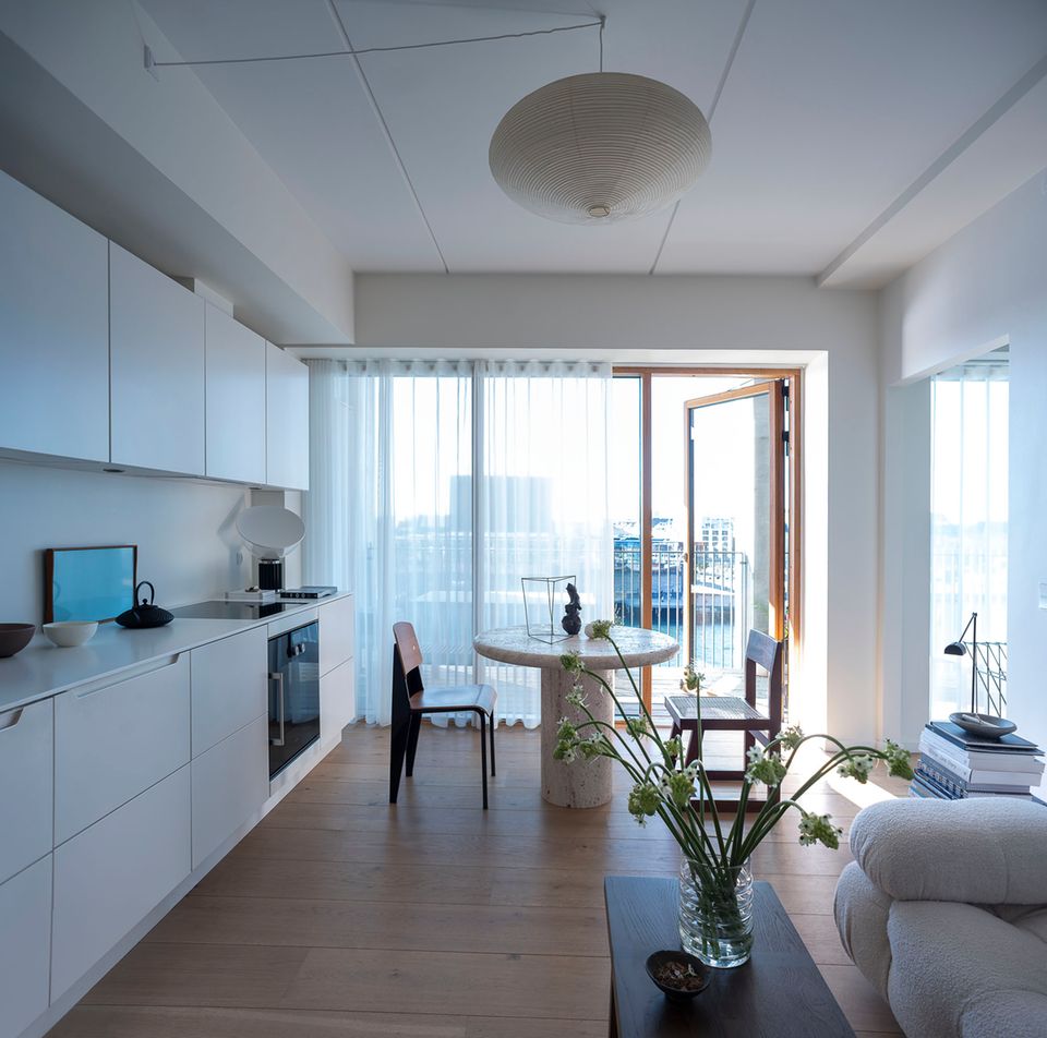 Offener Wohnbereich mit weißer Küche, rundem Esstisch und Blick über den Balkon aufs Wasser