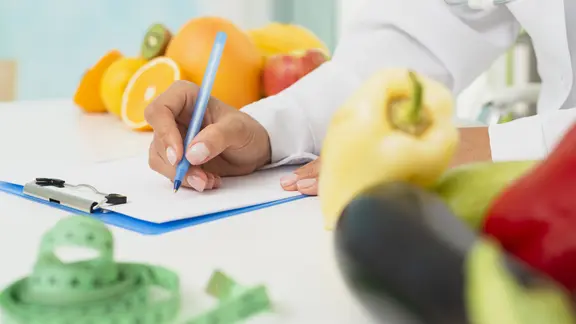 Stoffwechseldiät, Symbolbild: Person mit Arztkittel schreibt auf eine Kladde, daneben liegen ein Maßband, Obst und Gemüse. | IMAGO/ imageBROKER, Oleksandr Latk Stoffwechseldiät, Symbolbild: Person mit Arztkittel schreibt auf eine Kladde, daneben liegen ein Maßband, Obst und Gemüse.