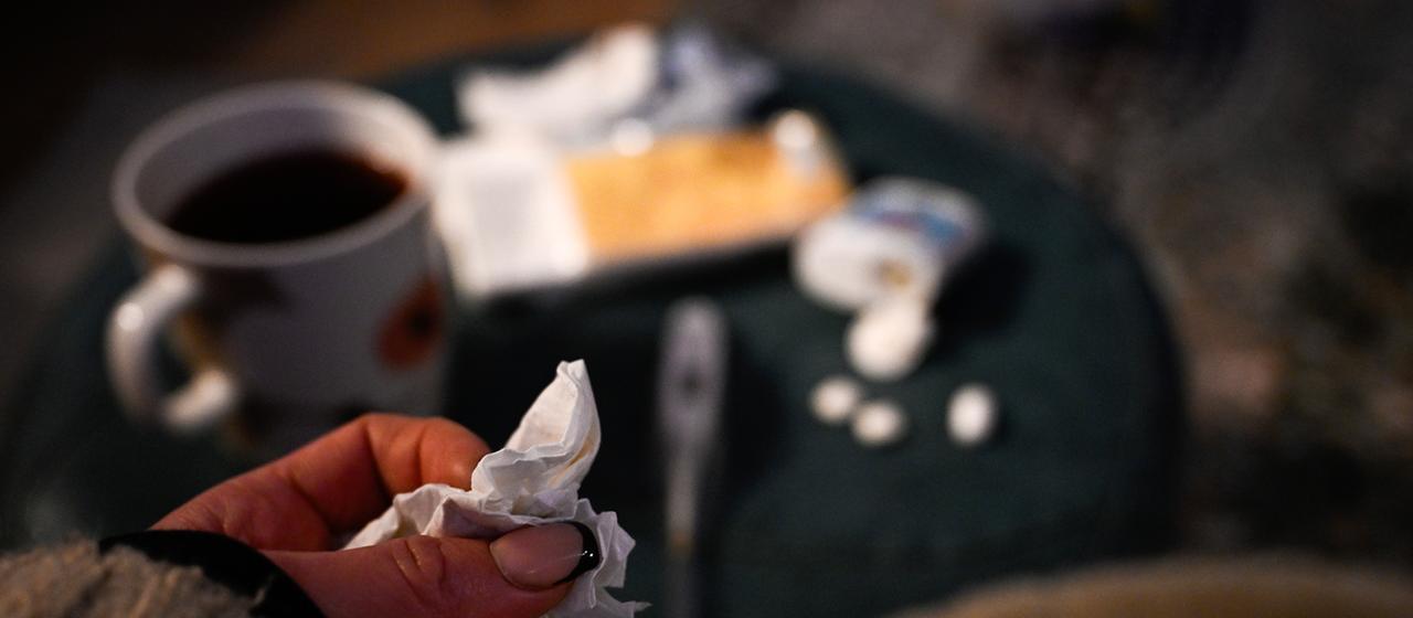 Eine Hand mit einem Taschentuch ist vor einem Tisch mit Tee, Fieberthermometer und Medikamenten zu sehen. | dpa Eine Hand mit einem Taschentuch ist vor einem Tisch mit Tee, Fieberthermometer und Medikamenten zu sehen.