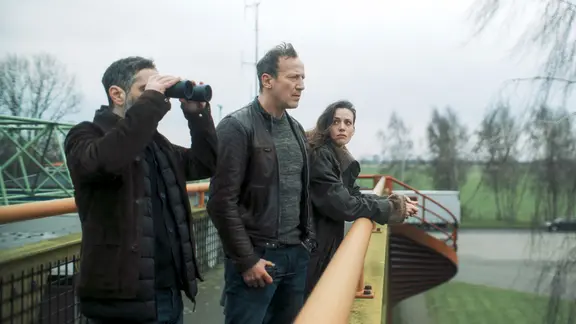 Mario Schmitt (Denis Moschitto, l.) Falke (Wotan Wilke Möhring, M.) und Lynn de Baer (Gaite Jansen) stehen auf einer Brücke.